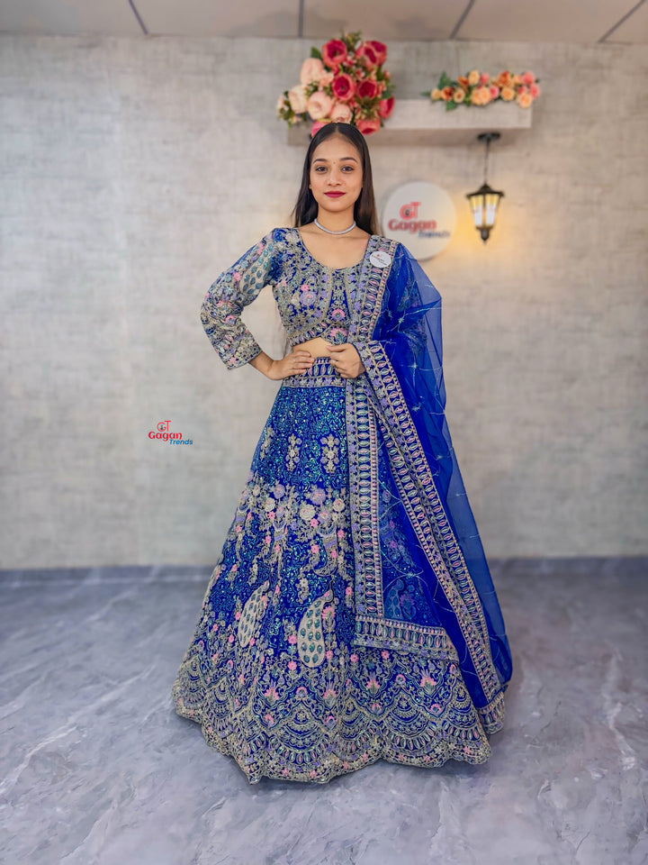 Woman in a blue embroidered traditional outfit standing against a light gray wall with floral decorations.